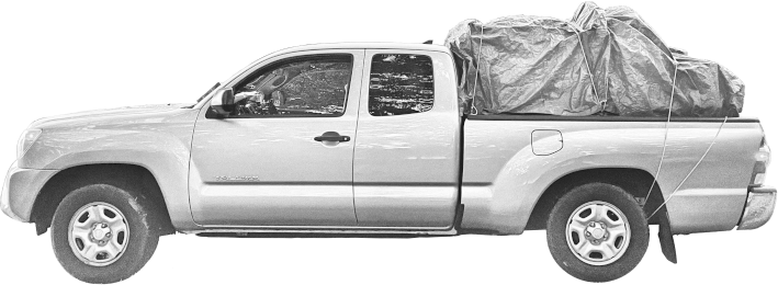 A greyscale pickup truck with a load of things in the back covered by a lumpy tarp