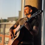 Patrick Kozub, standing, leaning against a concrete pillar, holding his guitar with the sunset on his face, partially obscured in a shadow
