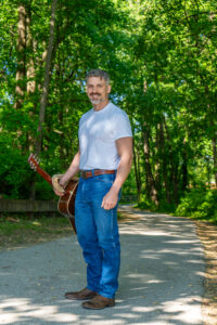 Patrick Kozub, a man holding a guitar in one hand and smiling