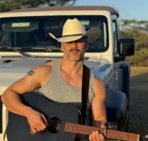 Patrick Kozub, a man, seated on the bumper of a Jeep holding a guitar